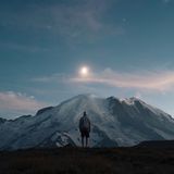 A figure in a hoodie stands facing a snow-capped mountain under a moonlit sky, with soft clouds and stars visible in the night.