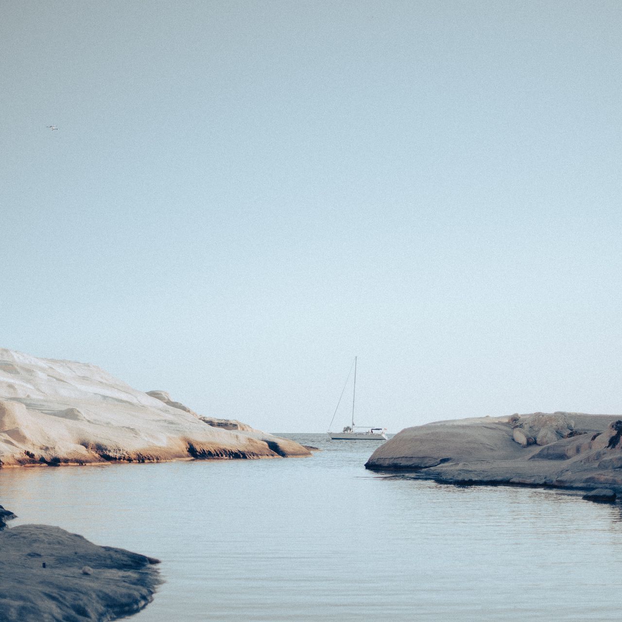 A serene coastal scene featuring a calm body of water between smooth, pale rocks. In the distance, a sailboat is anchored on the horizon under a clear sky.