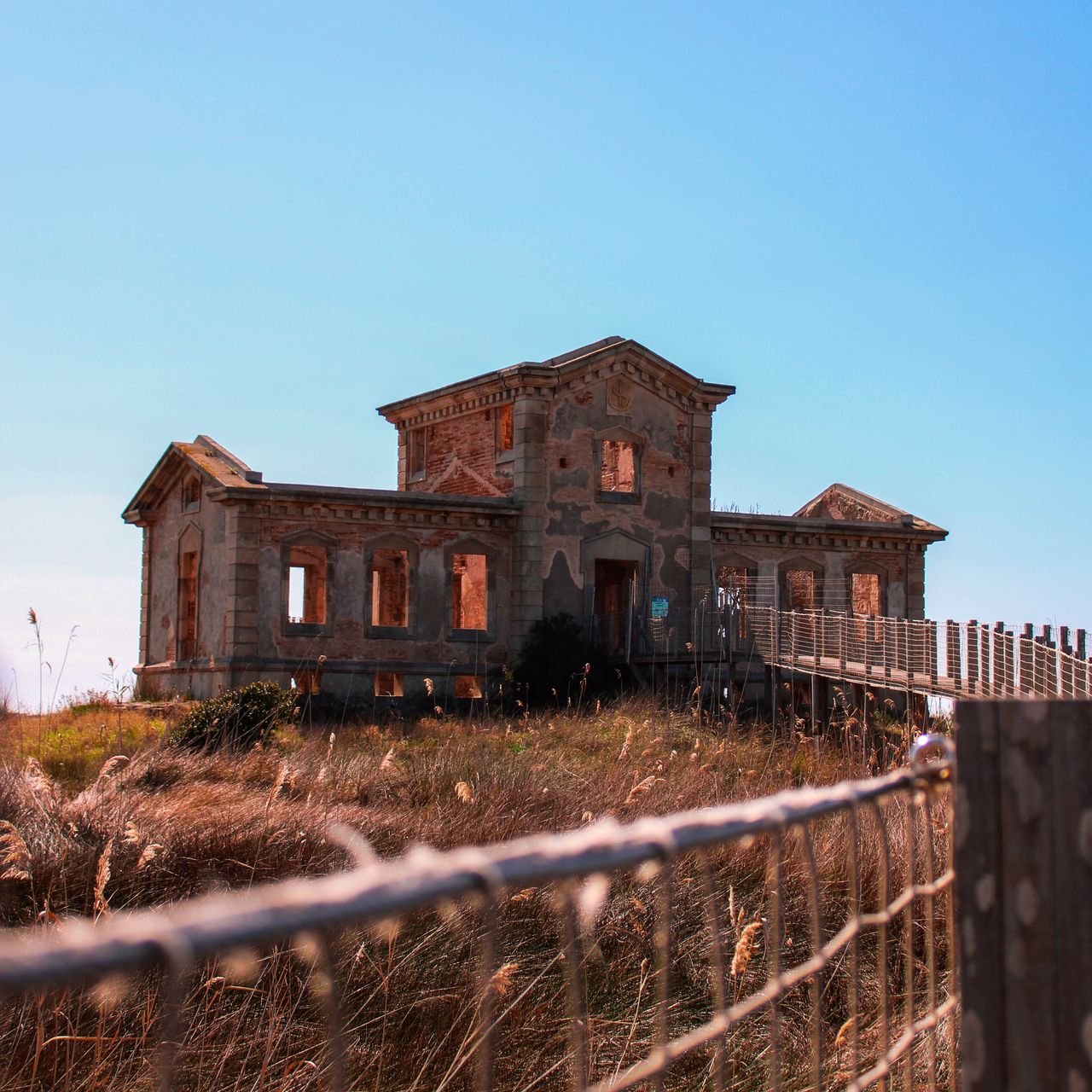 A weathered, two-story building with broken windows and a crumbling facade, surrounded by tall grasses and a wooden fence, set against a clear blue sky.