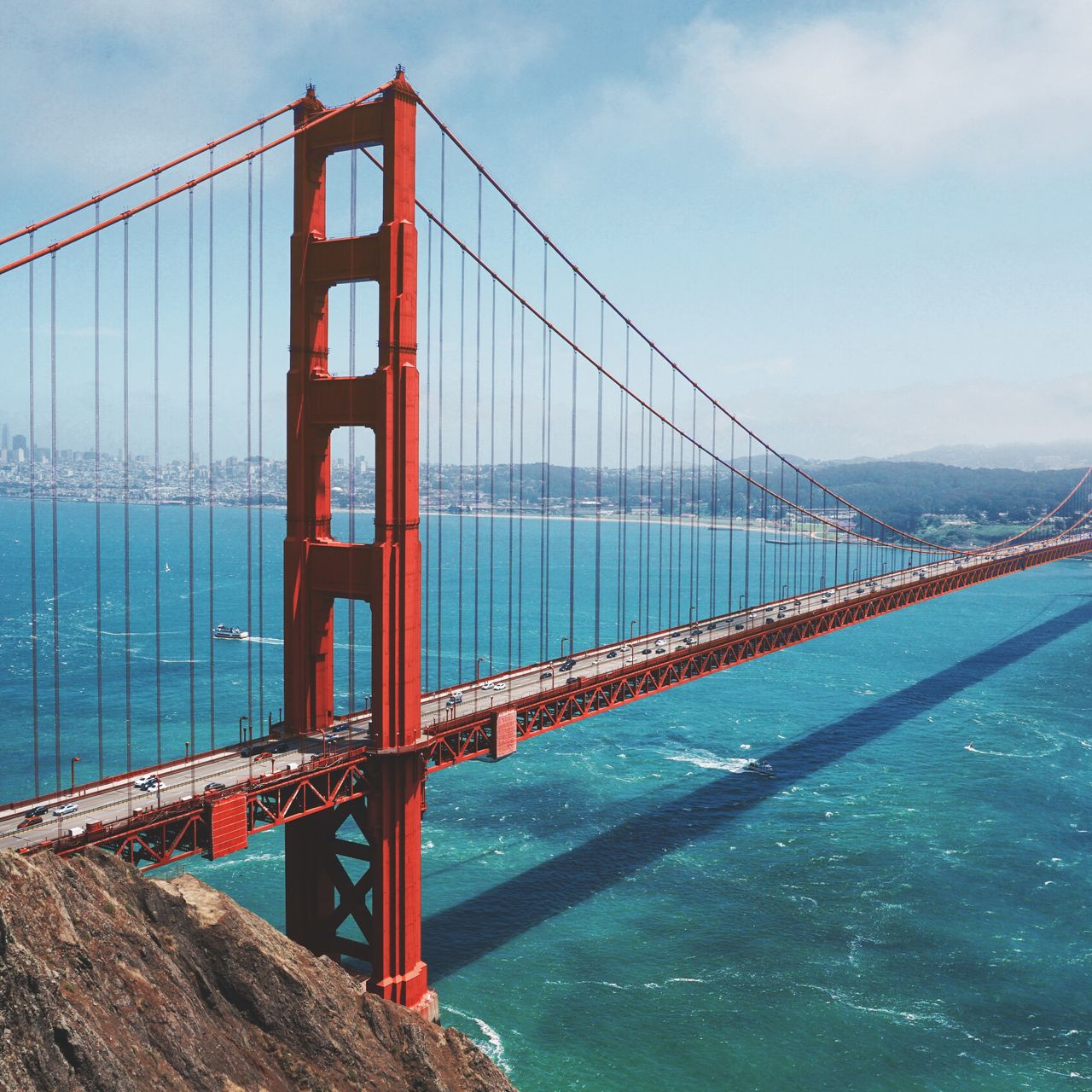 Aerial view of the Golden Gate Bridge spanning over turquoise water, with a city skyline visible in the background under a clear blue sky.