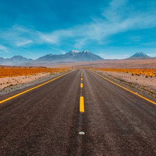 A straight road stretching towards distant mountains under a clear blue sky, with desert terrain and sparse vegetation on either side.