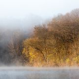 A foggy scene featuring a tranquil lake bordered by trees with bare branches and hints of yellow and red foliage in the background. The water reflects the surrounding mist and landscape.