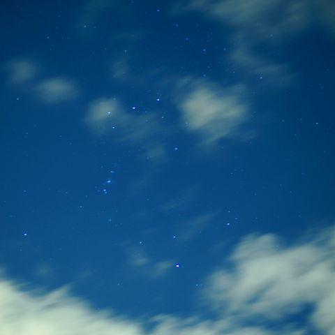 A night sky filled with stars, partially obscured by soft, wispy clouds. The background is a deep blue, with various stars shining brightly.