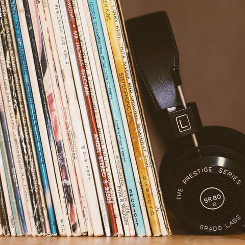 A close-up of a stack of vinyl records with a pair of black Grado Labs headphones resting against them. The records feature various title labels in a mix of colors.