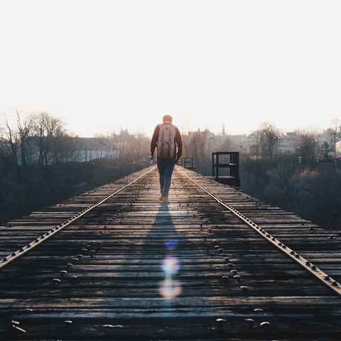 A lone figure walks along a wooden train trestle, with a backpack, towards the horizon. The scene is bathed in soft, warm light, surrounded by bare trees and distant buildings.