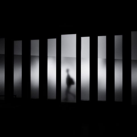 A silhouette of a person walking past frosted glass panels in a dimly lit space, creating a stark contrast with a dark background.