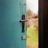 Image of a partially open turquoise door with a decorative handle, revealing a blurred green background.