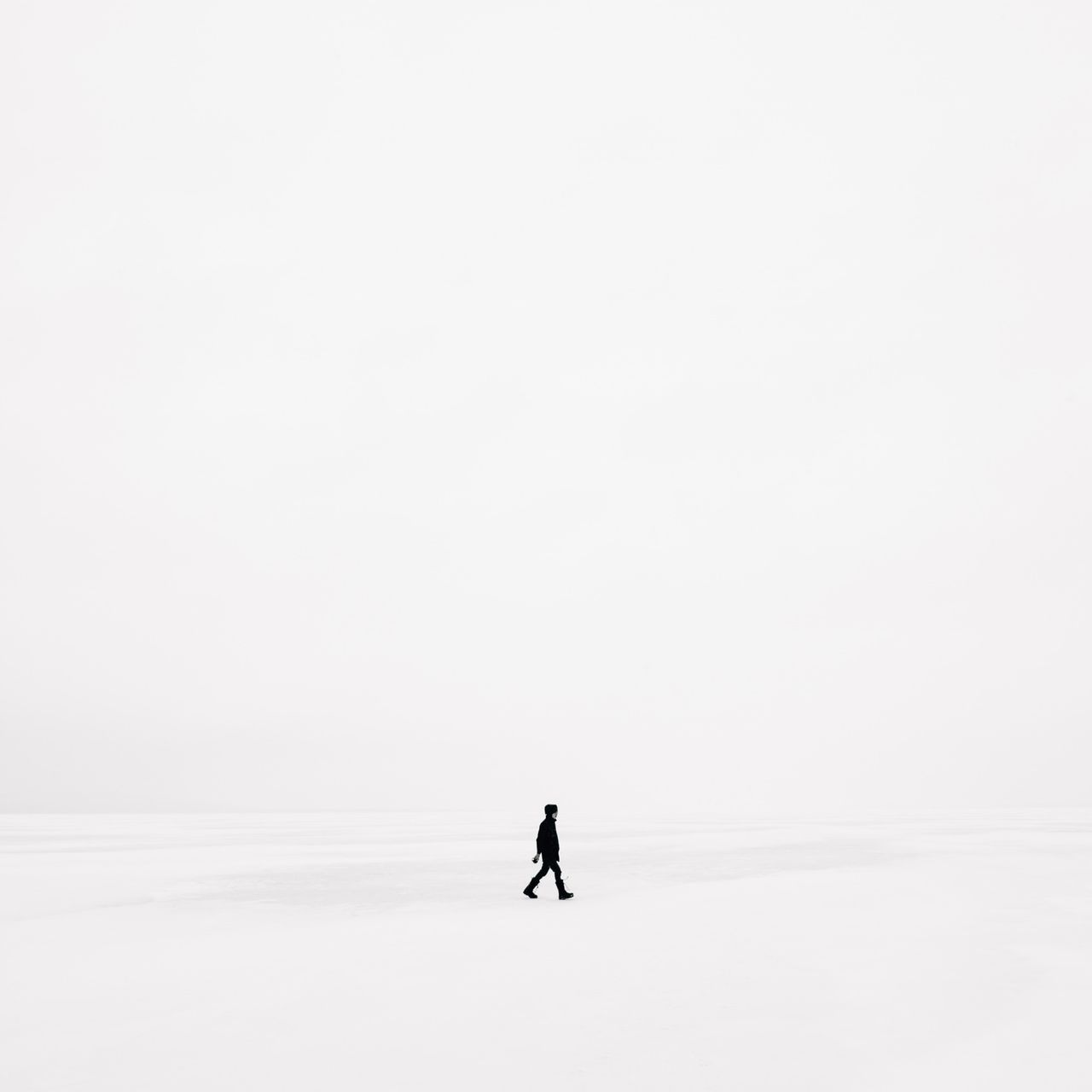 A solitary figure in black clothing walks across a vast, white, snowy landscape, under an overcast sky.