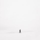 A solitary figure in black clothing walks across a vast, white, snowy landscape, under an overcast sky.