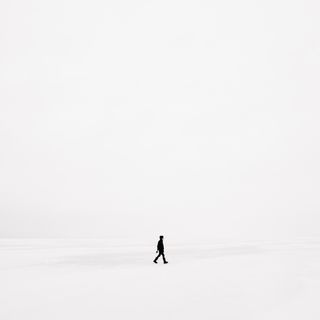 A solitary figure in black clothing walks across a vast, white, snowy landscape, under an overcast sky.