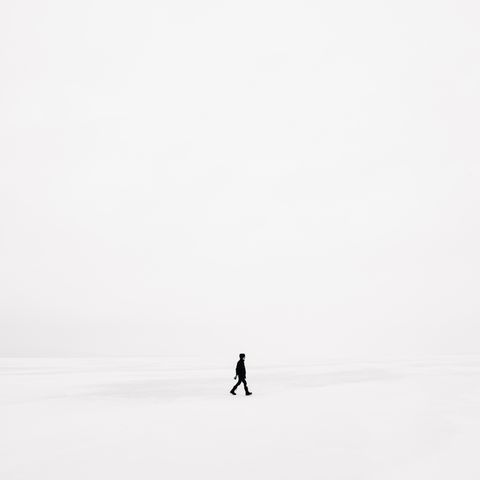 A solitary figure in black clothing walks across a vast, white, snowy landscape, under an overcast sky.